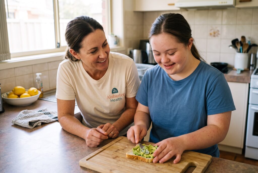 An Alabaster Health support worker crouches beside a teenage girl with Down syndrome, encouraging her as she makes her own lunch in a home kitchen.